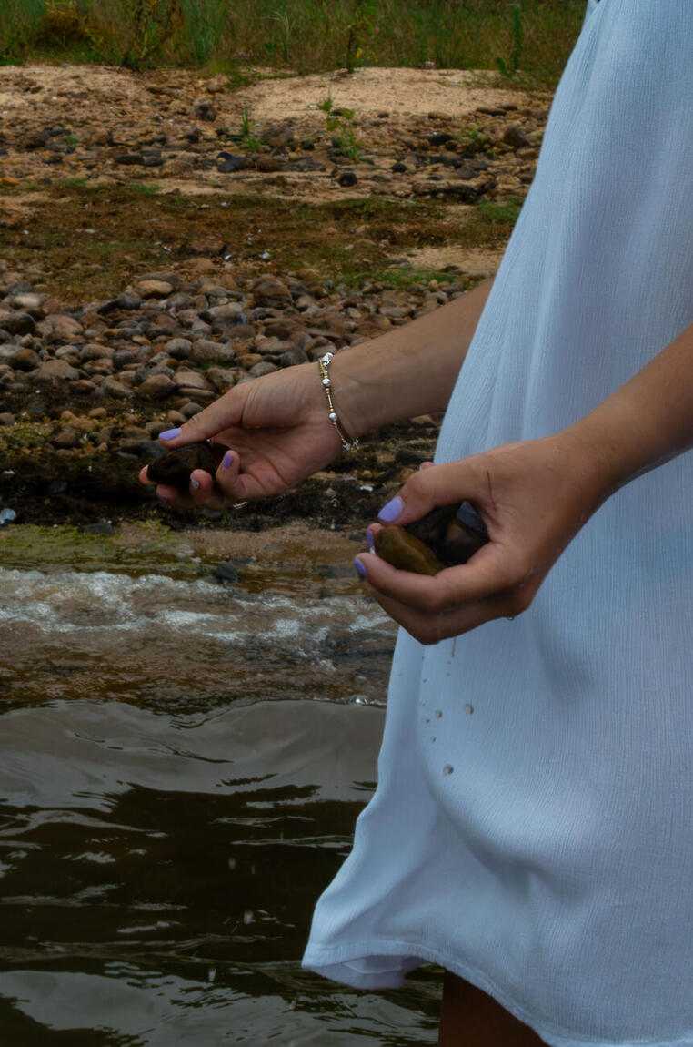 Two hands holding wet stones against a white dress at the shoreline