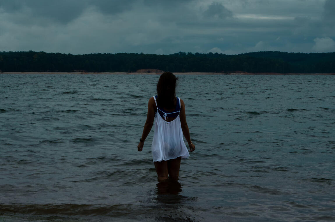 A figure walking away from the camera into open water at dusk